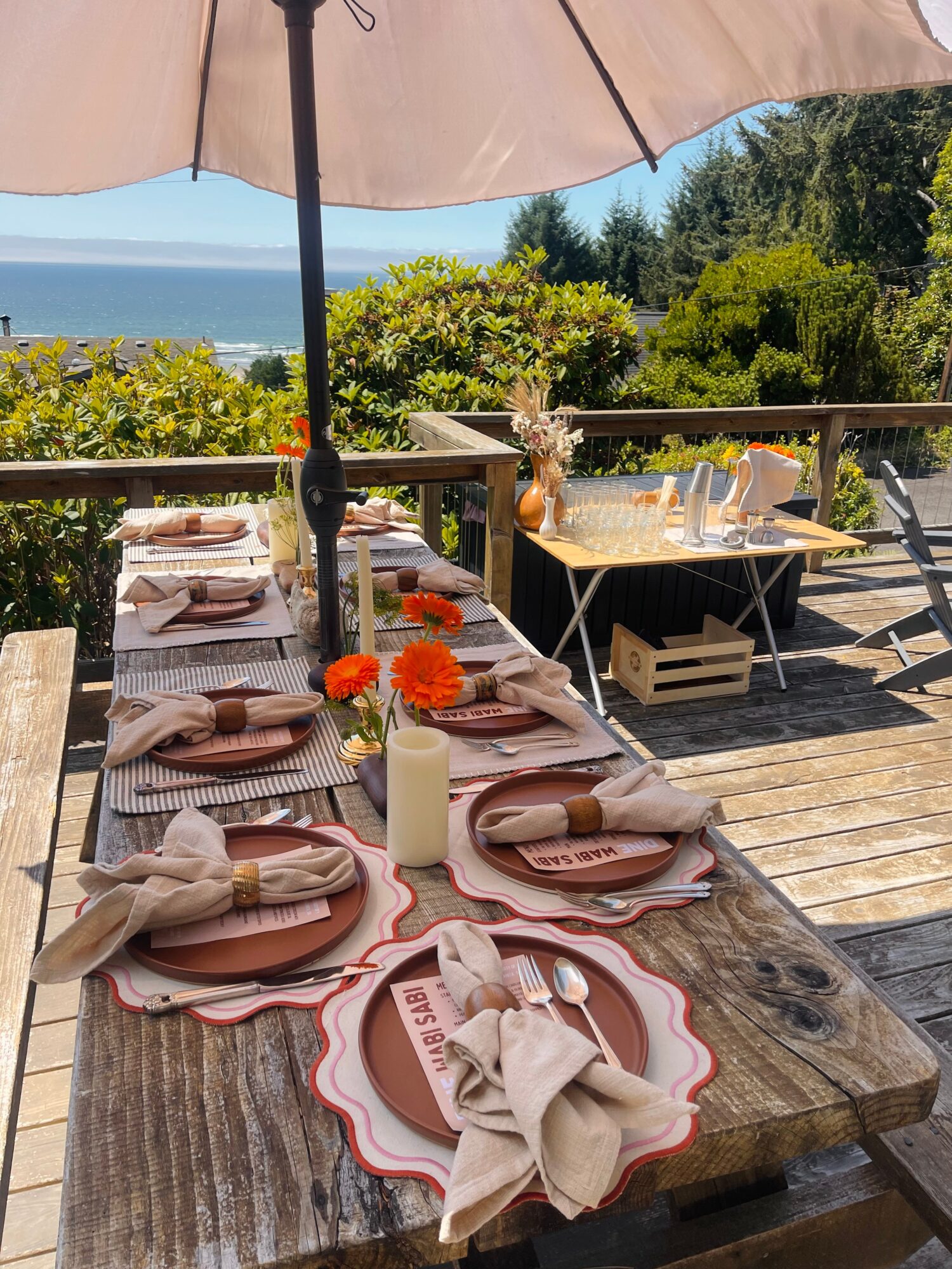 Outdoor dining table set with plates, folded napkins, orange flowers, and an umbrella, overlooking greenery and the ocean.