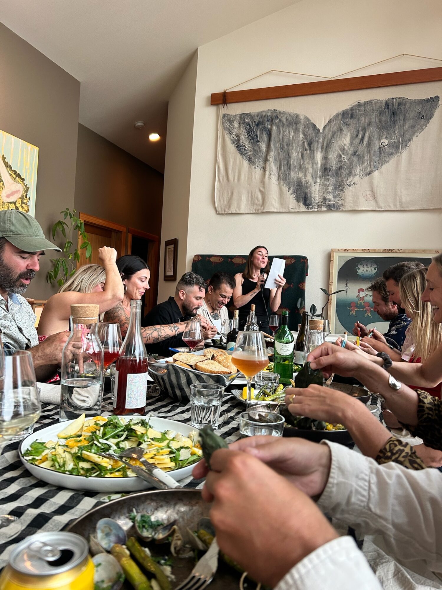 People gathered around a dining table covered with dishes, drinks, and a striped tablecloth beneath a whale-tail wall hanging.