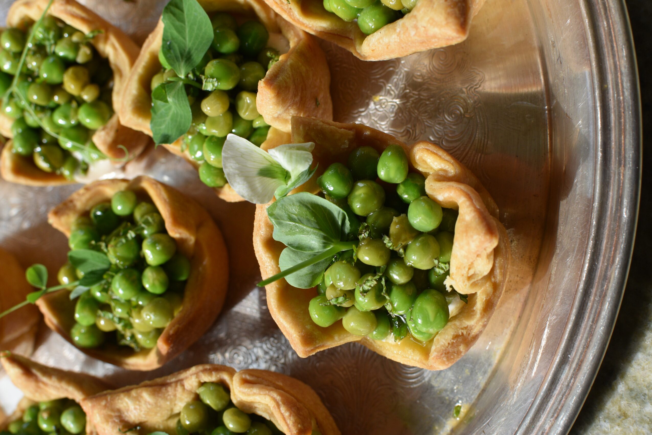 Crisp pastry cups filled with green peas and leafy herbs arranged on a round metal tray.