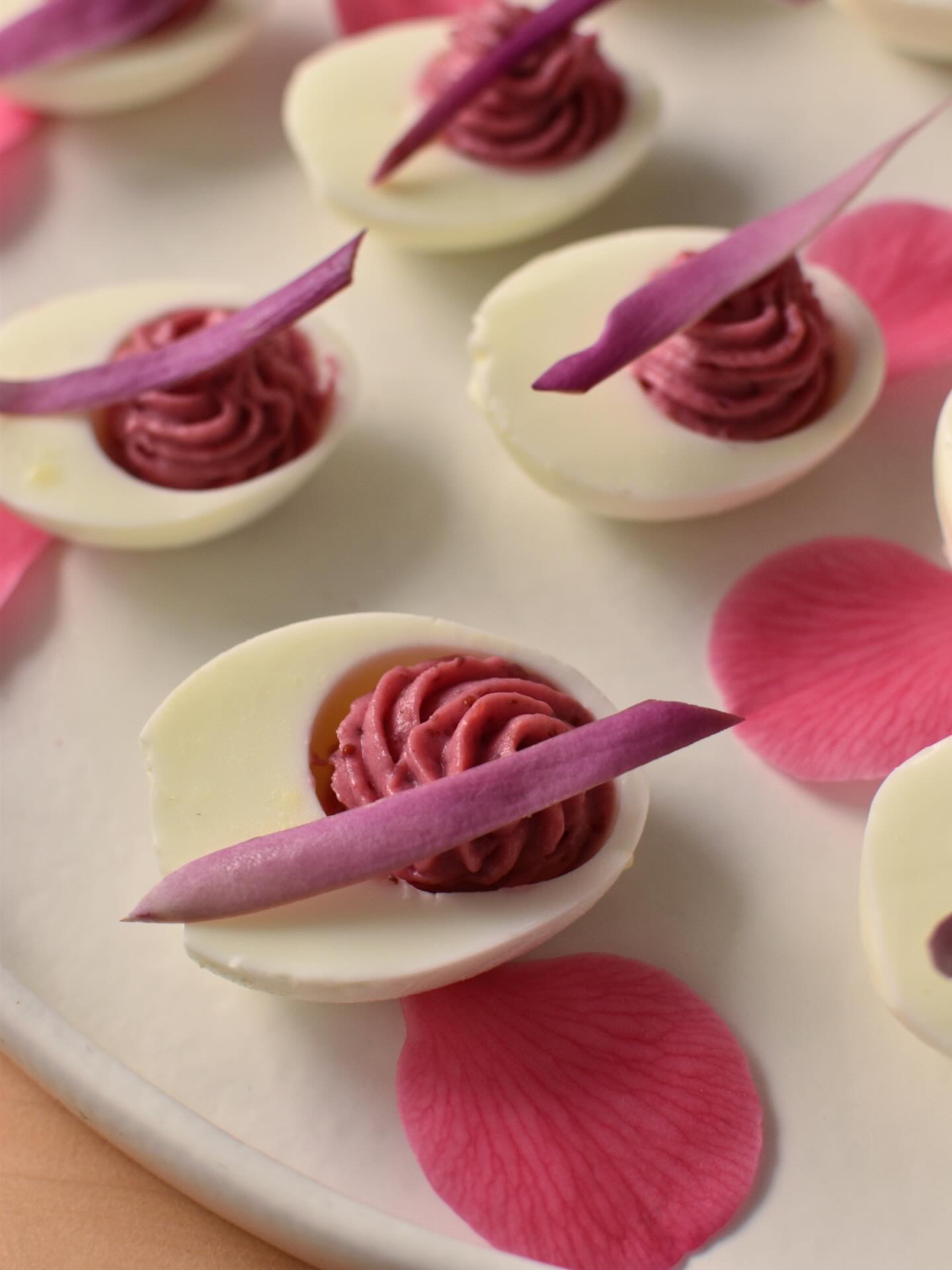 Deviled eggs topped with piped filling and purple petals, arranged on a white plate with pink flower petals.