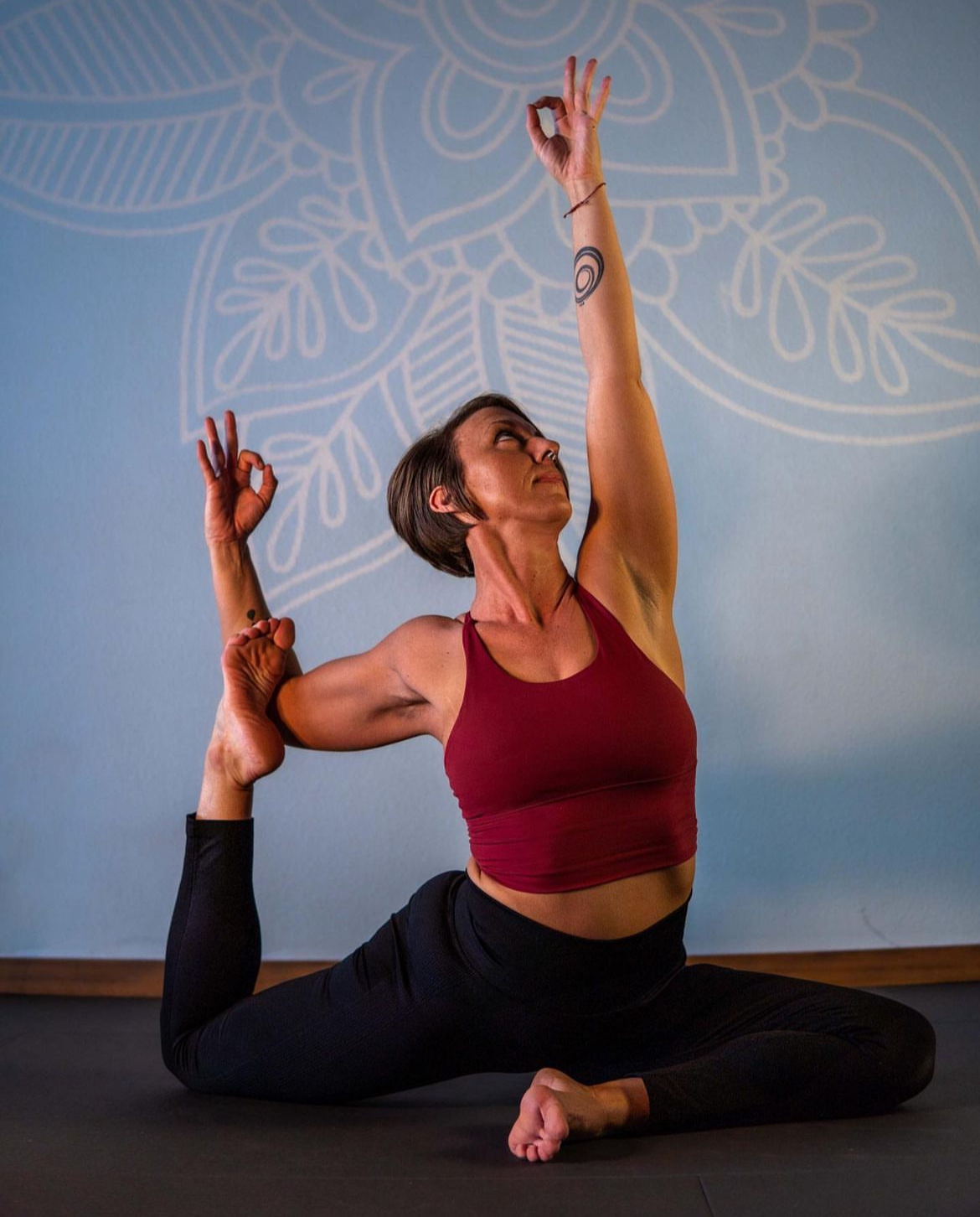 A person in a red top sits on the floor in a yoga pose, holding one foot and reaching an arm overhead.