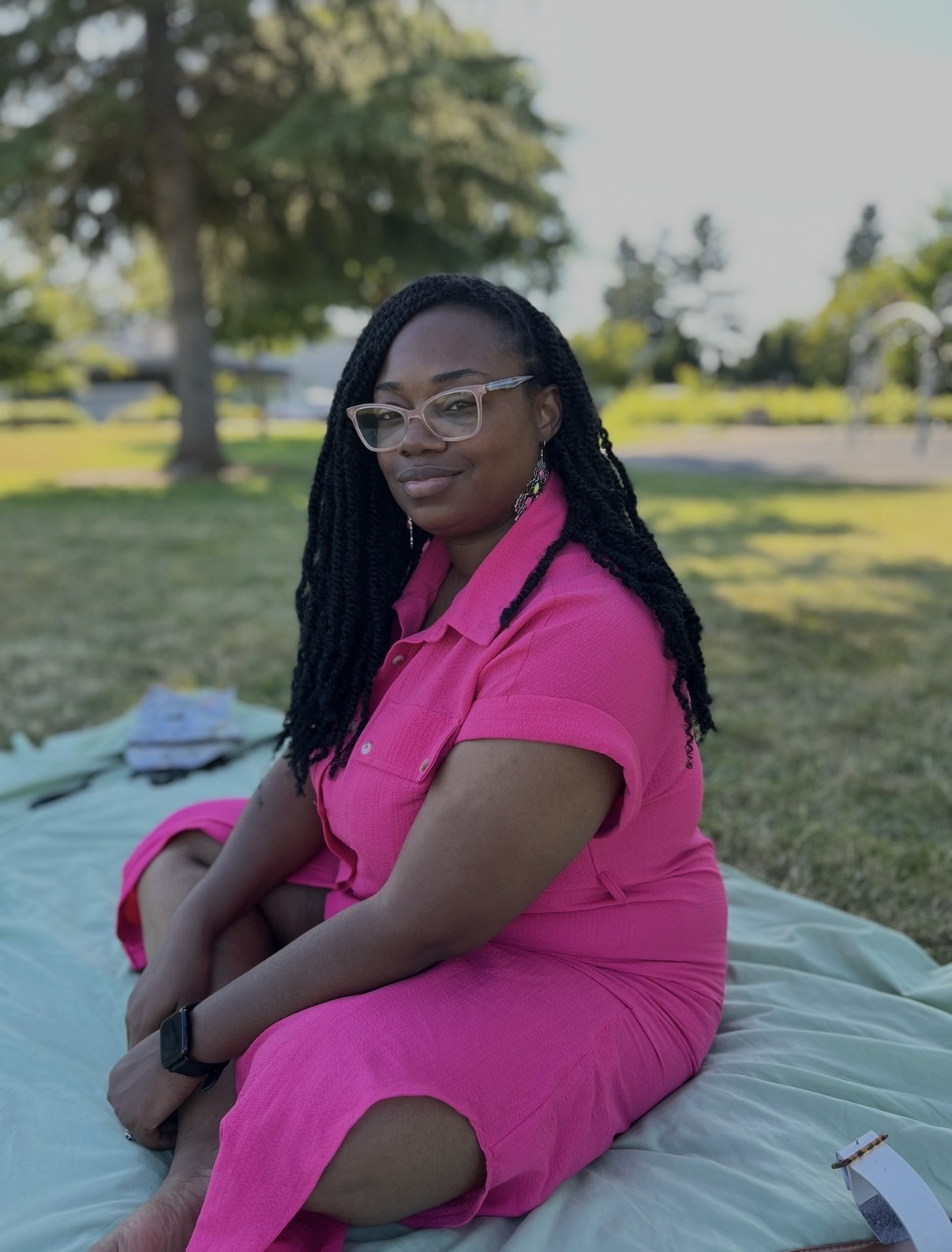 A person wearing glasses and a bright pink dress sits on a blanket in a grassy park.