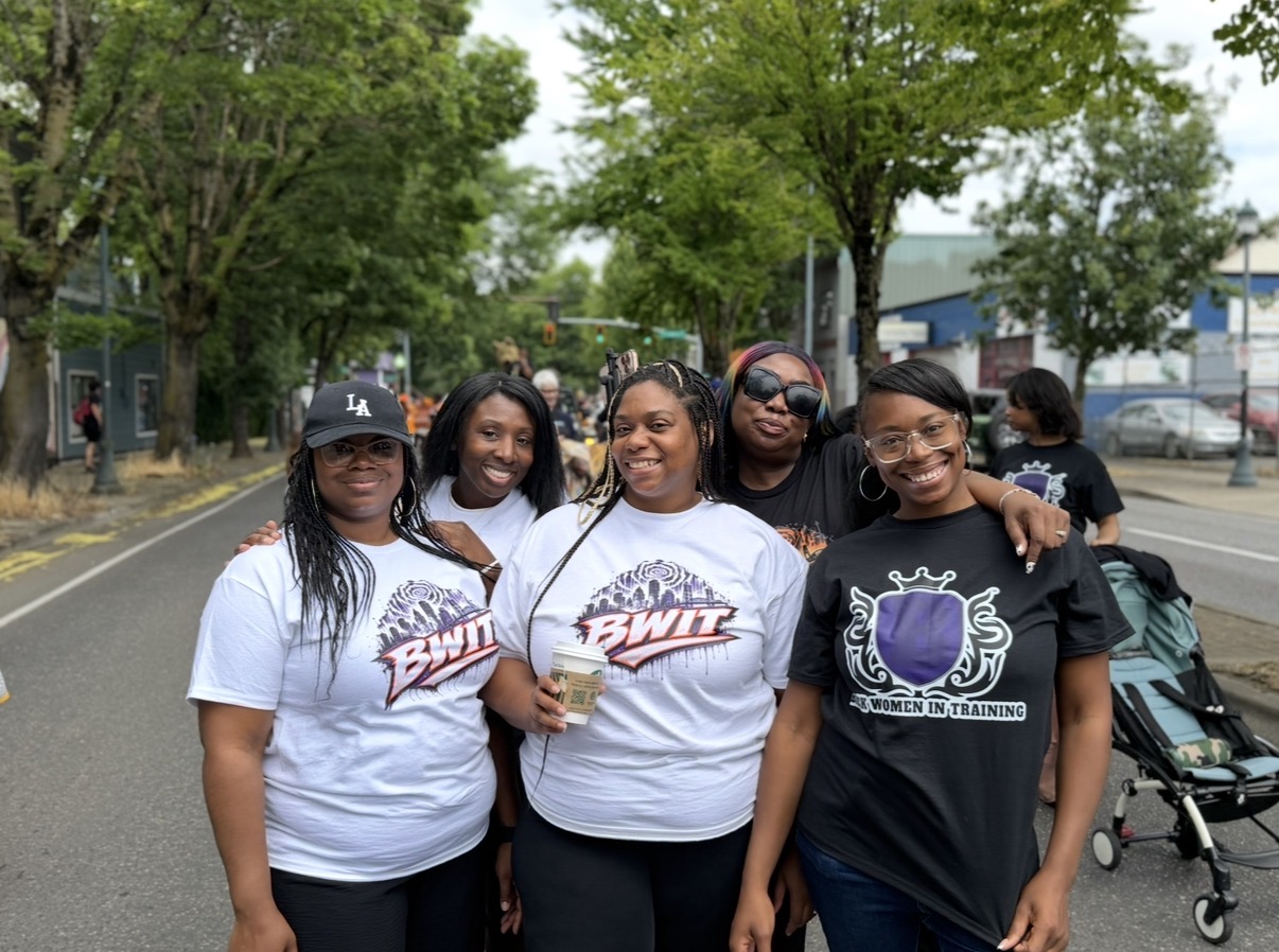 Five people pose together on a tree-lined street, wearing graphic T-shirts.