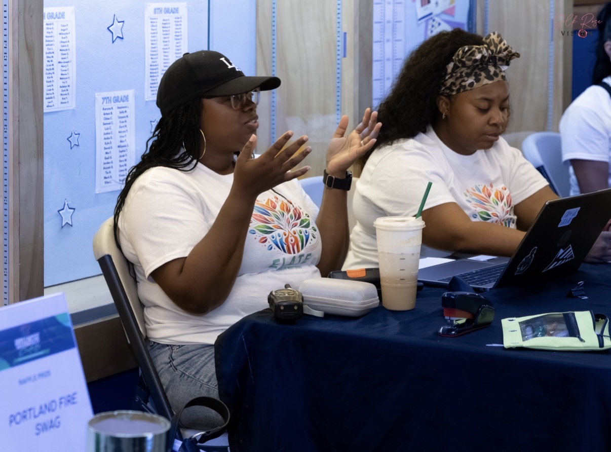 Two people sit at a table with laptops, a drink cup, and papers; one gestures while speaking.