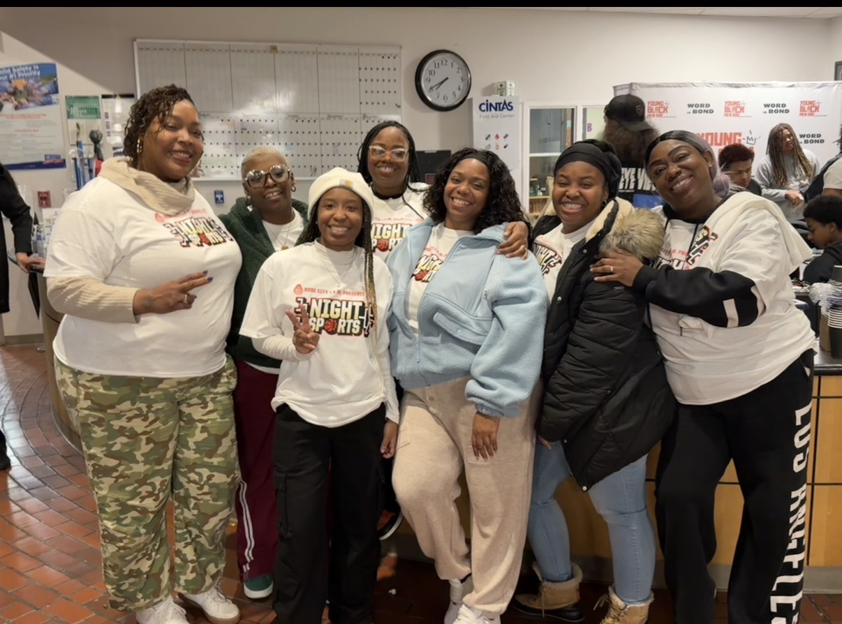 A group of people pose together in a classroom, with a whiteboard, wall clock, and tables behind them.