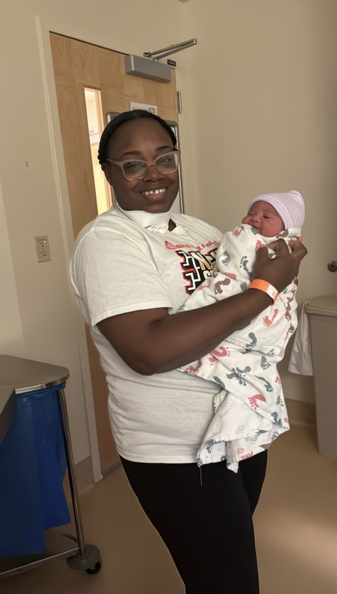 A person wearing glasses holds a newborn wrapped in a patterned blanket in a hospital room.