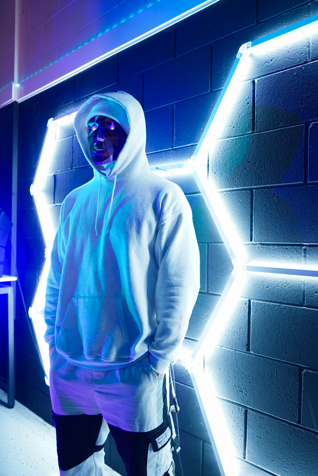 Person wearing a white hooded sweatshirt and shorts stands beside bright geometric lights on a dark brick wall.