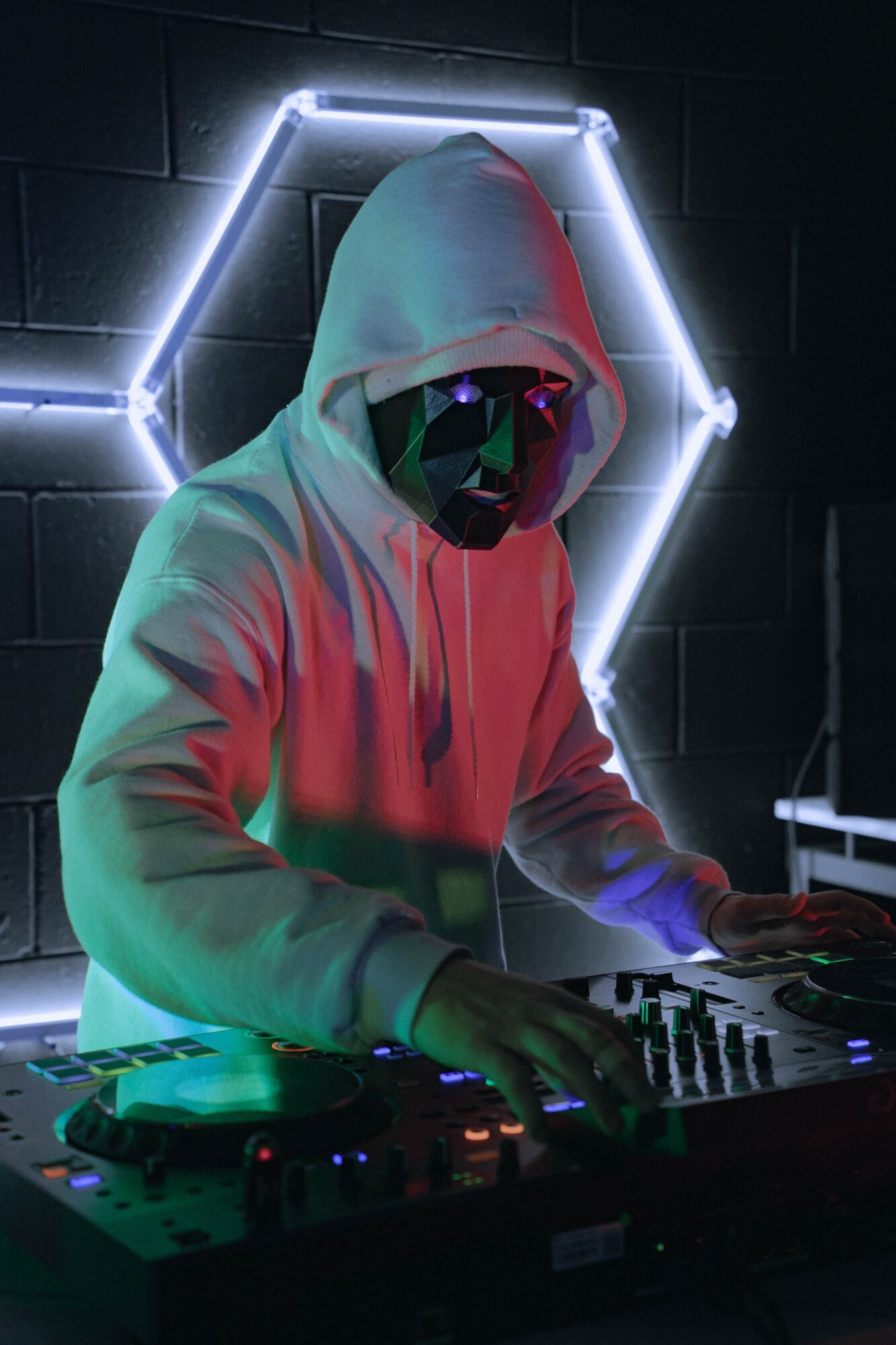 Hooded person wearing headphones operates a DJ controller in front of a glowing geometric light.