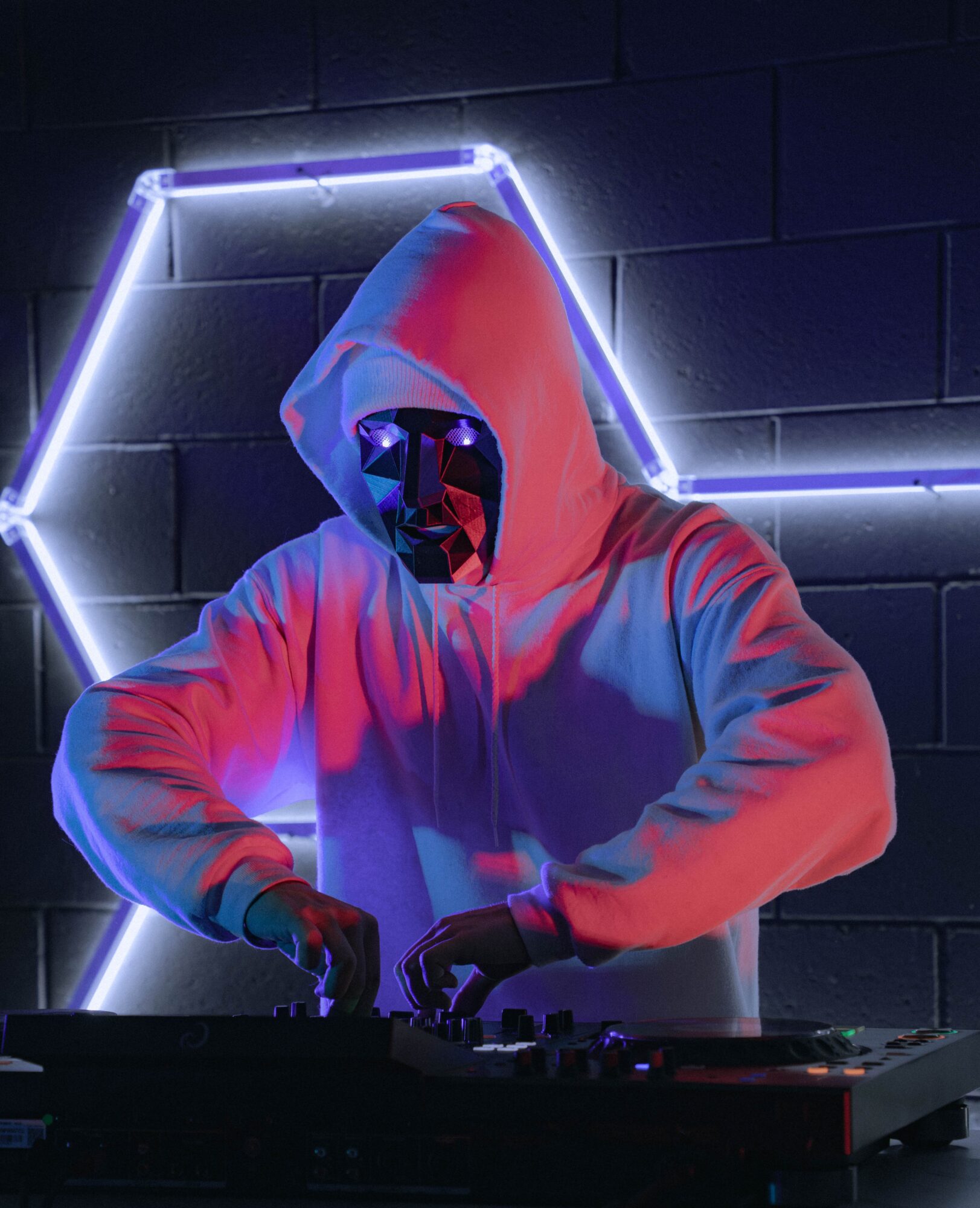 Hooded person wearing a reflective mask stands behind a DJ controller against a neon-lit brick wall.