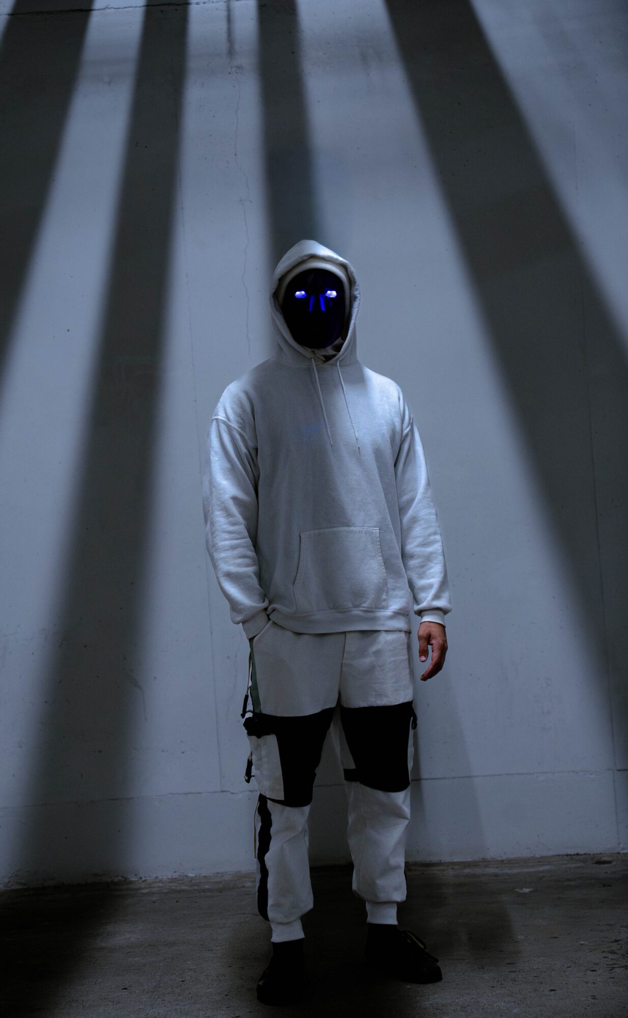 Person wearing a white hooded outfit and dark face covering stands beneath alternating light and dark vertical stripes.