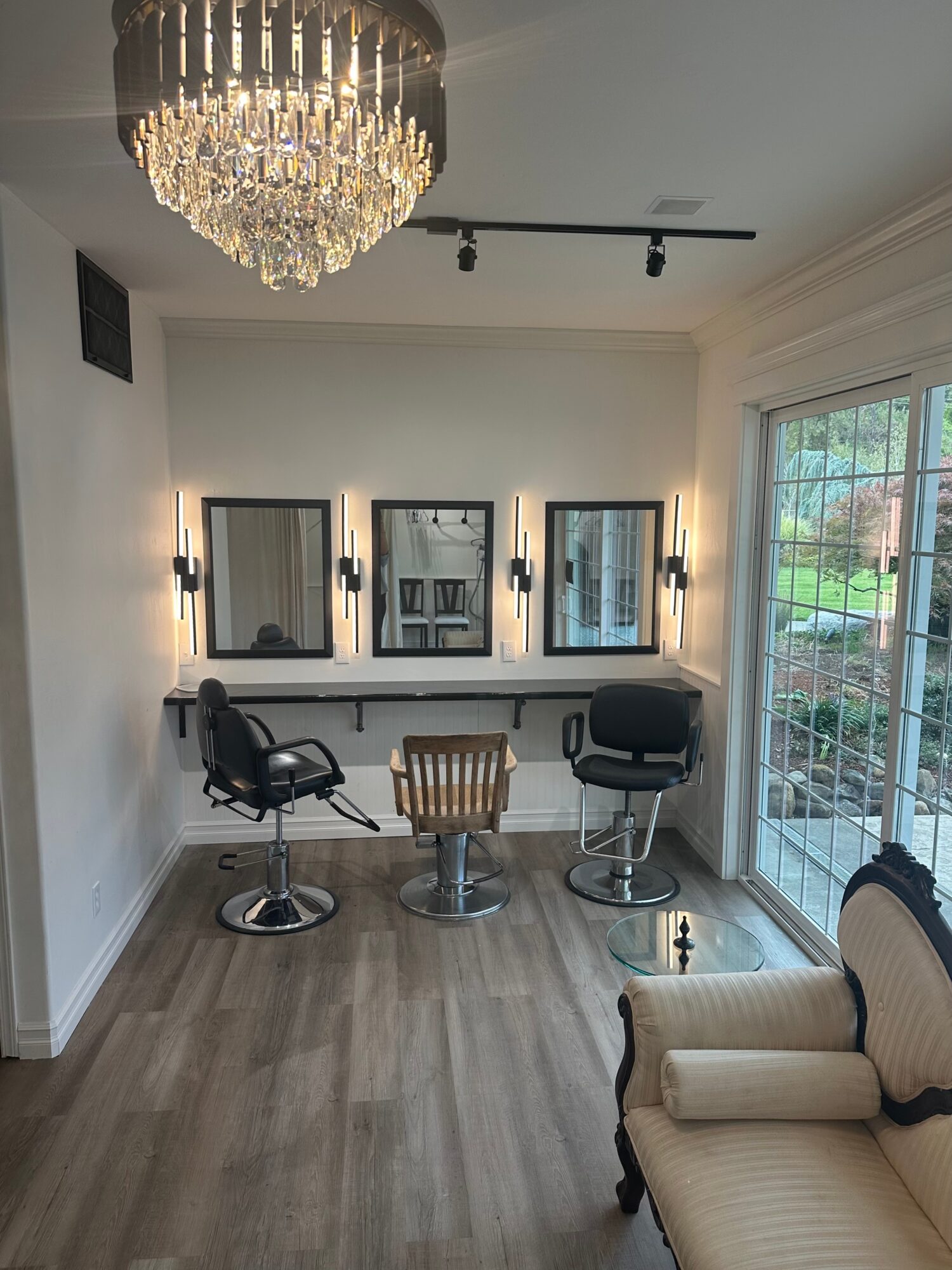 Bright salon with three styling stations, chairs, a chandelier, and large windows beside a cream sofa.