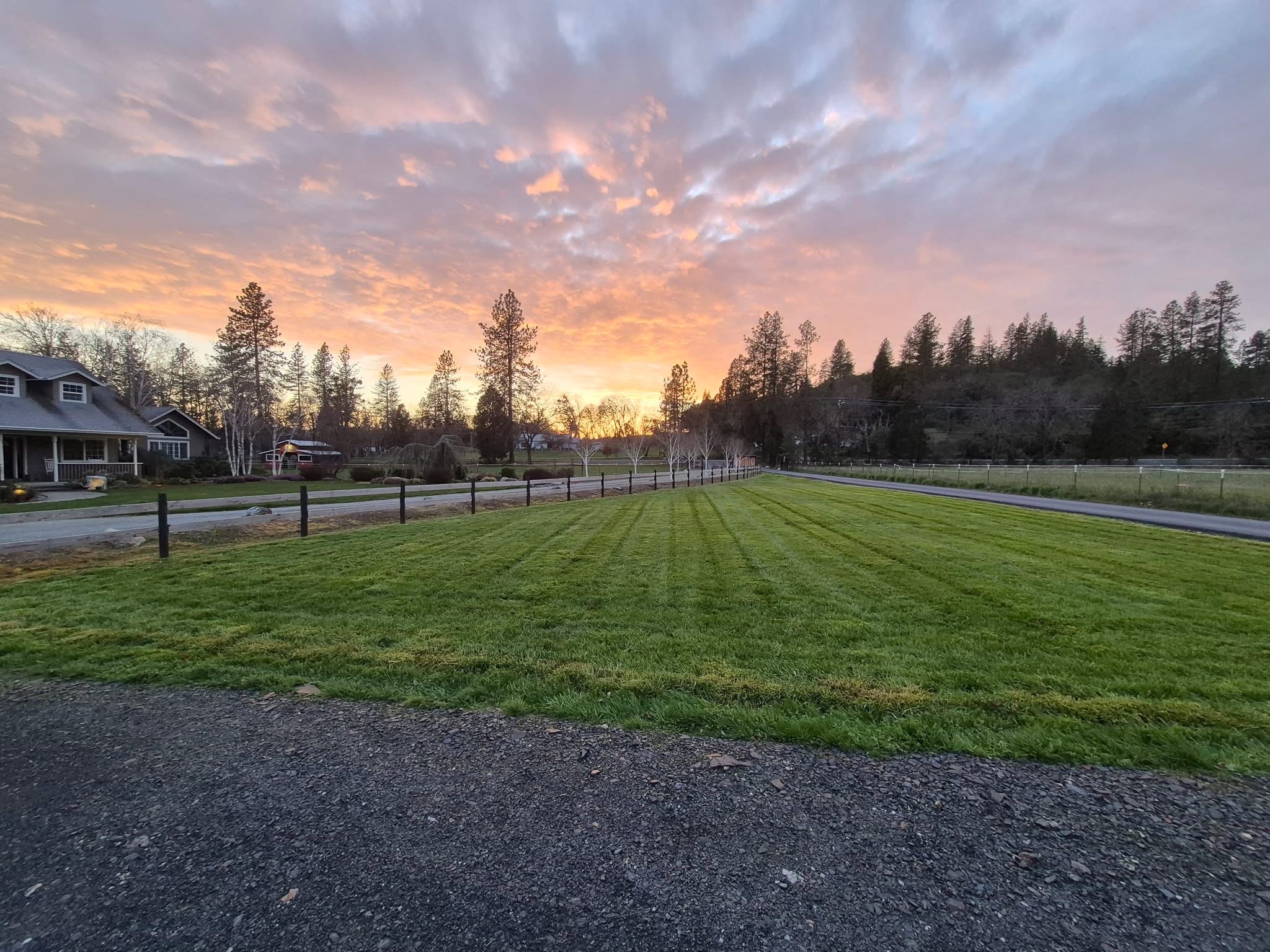 A grassy field and paved path beneath a colorful sunset sky, with houses and trees along the horizon.