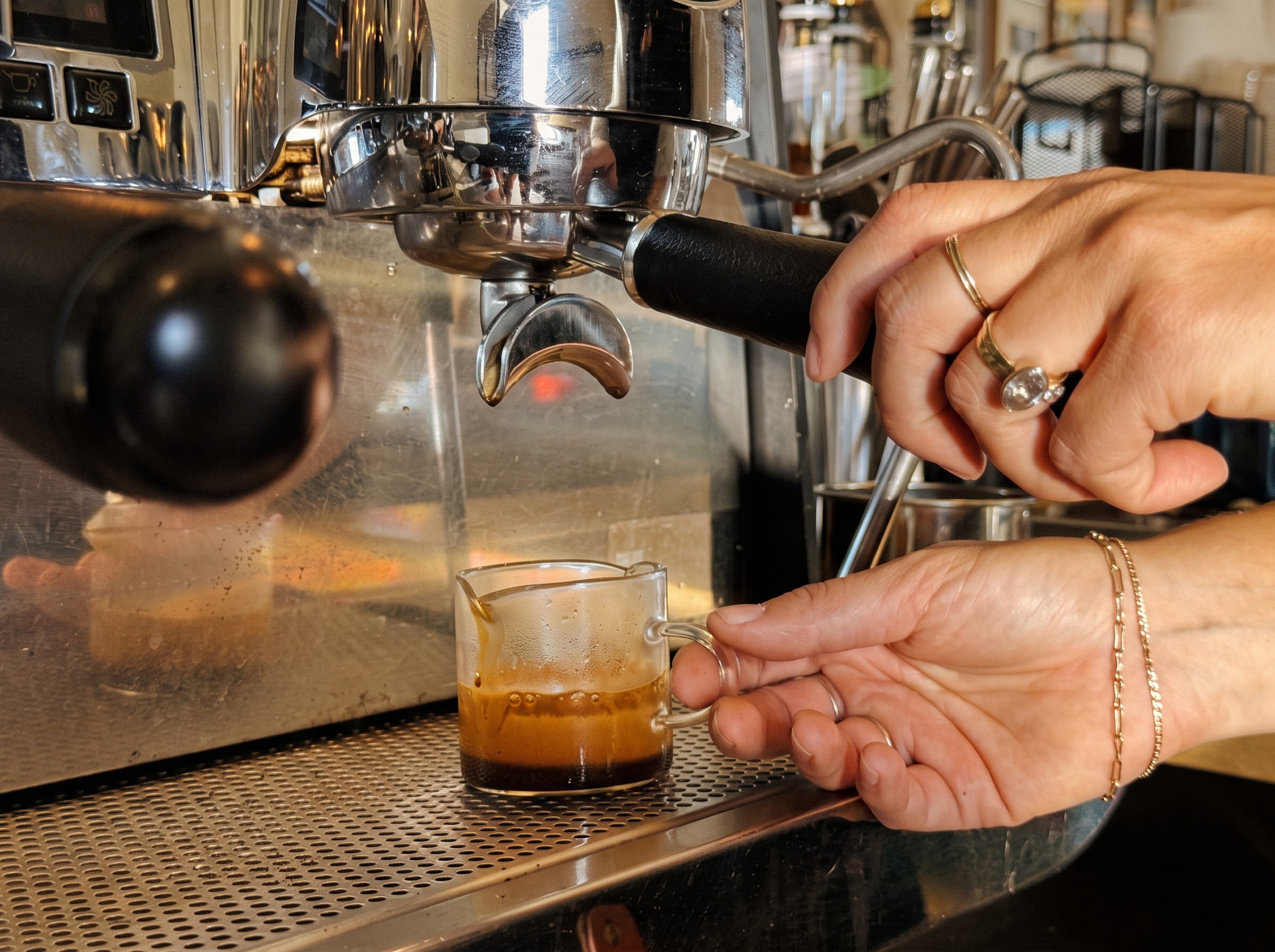 A hand holds an espresso machine portafilter while another steadies a small glass of espresso beneath the spout.