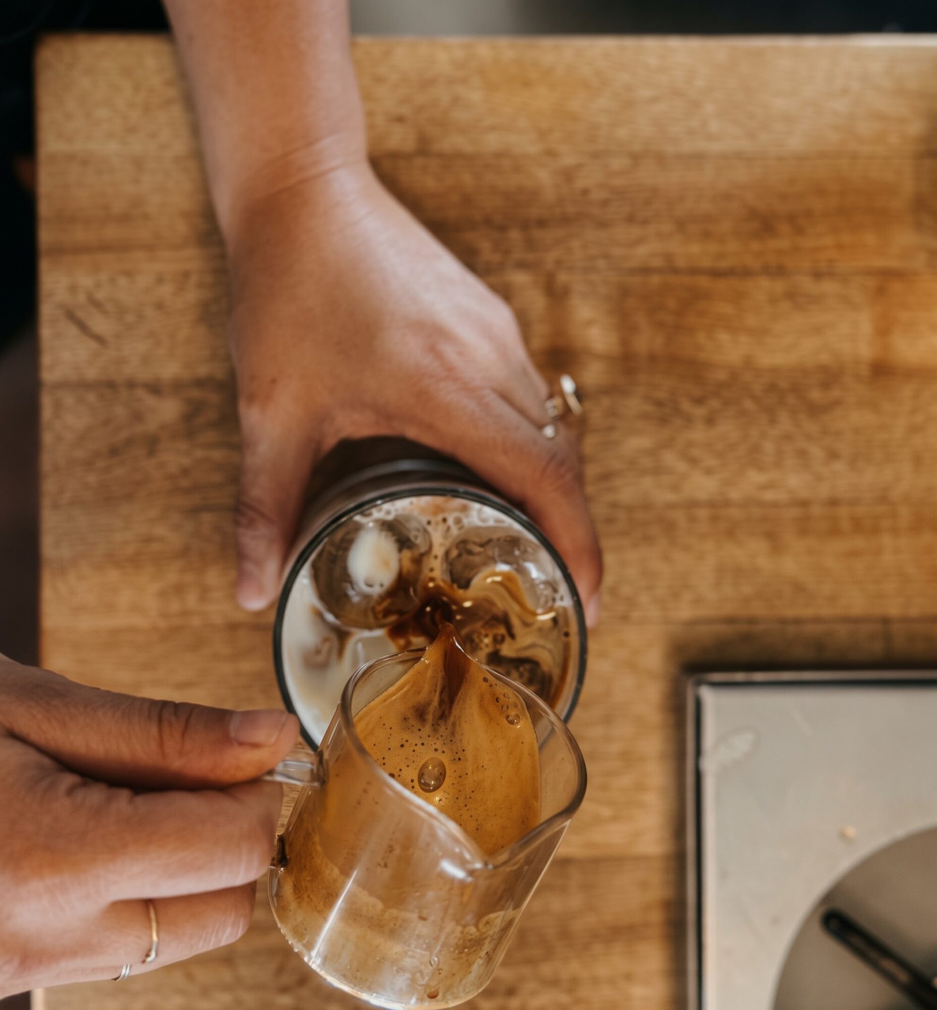 Hands pour a brown drink from a glass jar into a clear mug on a wooden surface.