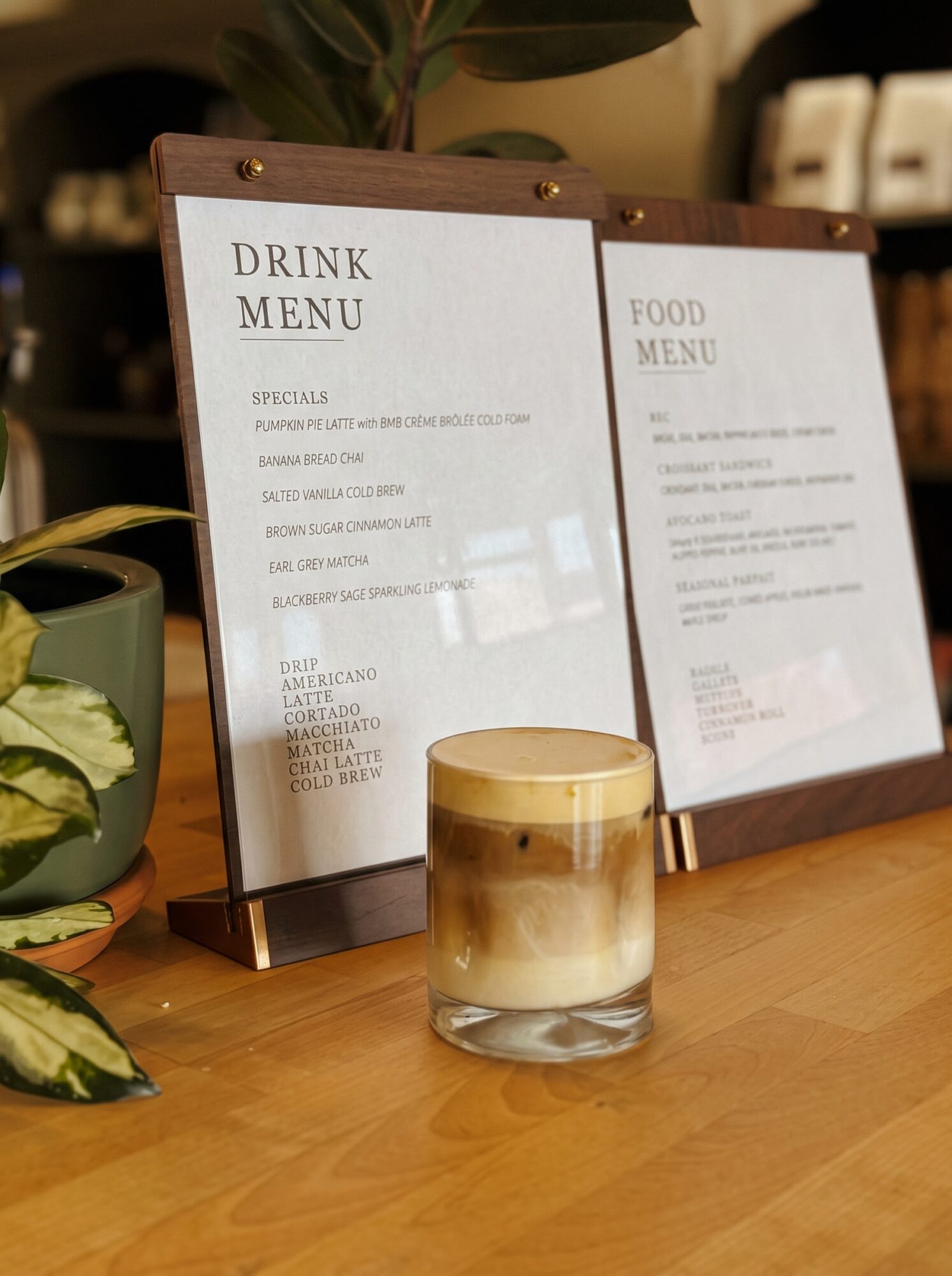 Two framed café menus stand on a wooden counter beside a layered candle and potted plants.