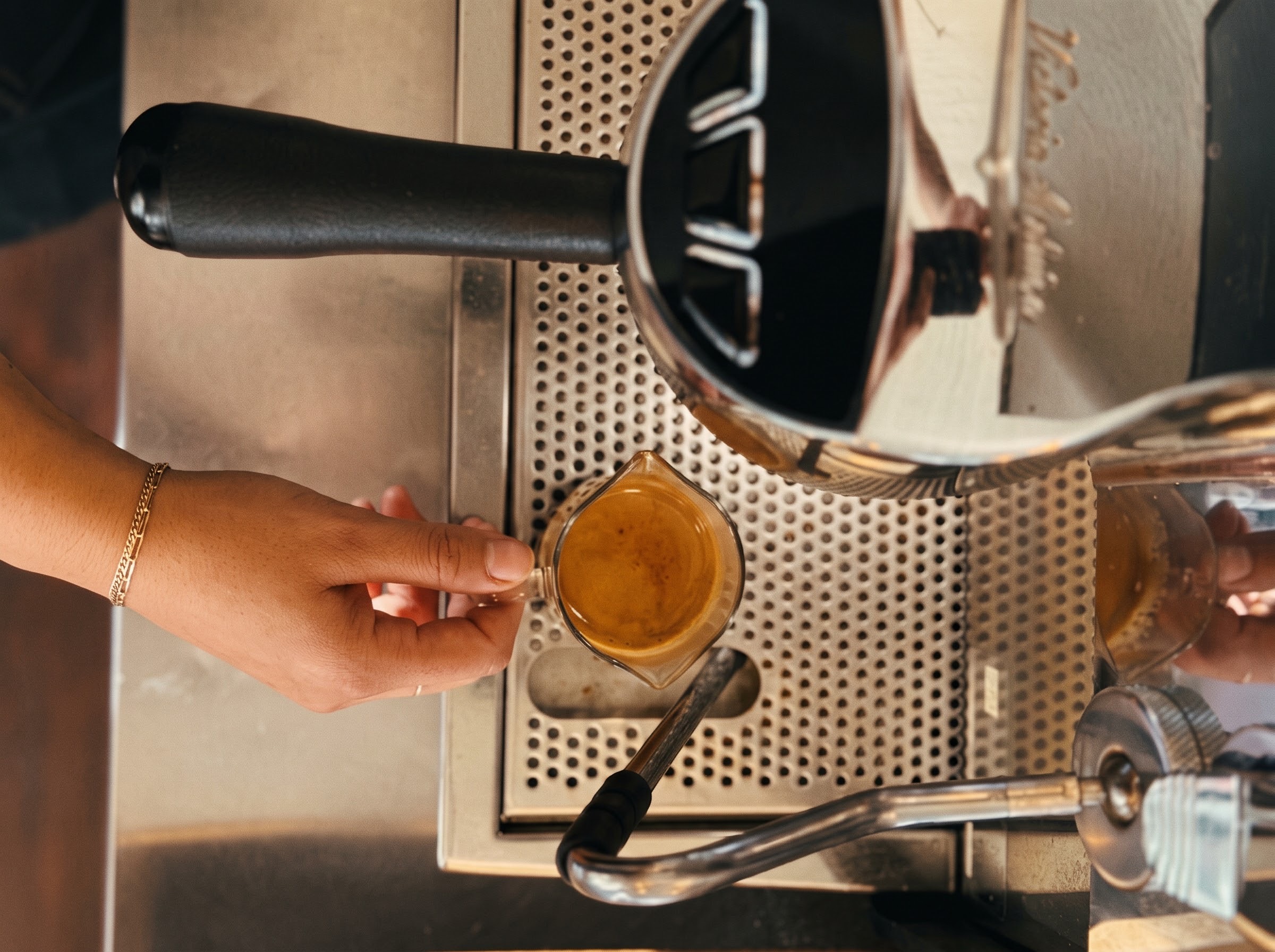 A hand holds a glass beneath a coffee machine spout, beside a metal drip tray and steam wand.